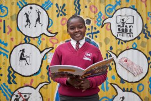 A girl wearing burgundy school uniform smiles whilst holding a book in Kibera, Kenya.