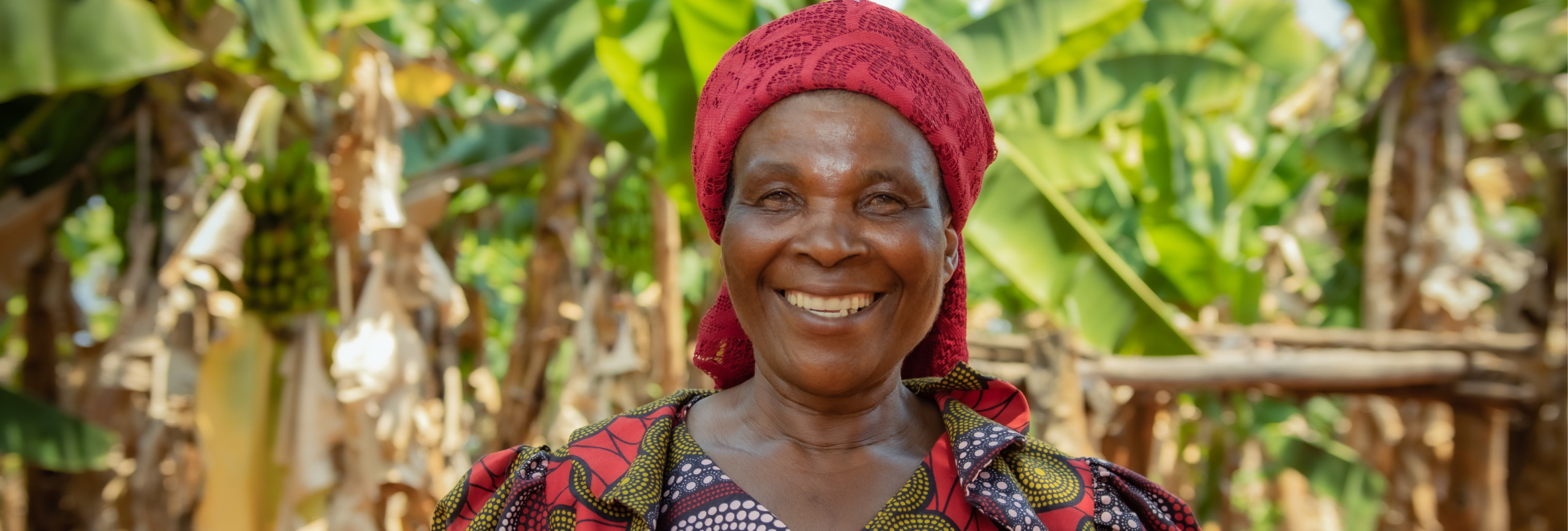 A woman wearing a red headscarf and patterned dress smiles at the camera in front of a green leafy backdrop.