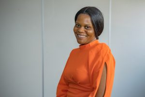 Tapiwa wears an orange dress and is smiling against a white background