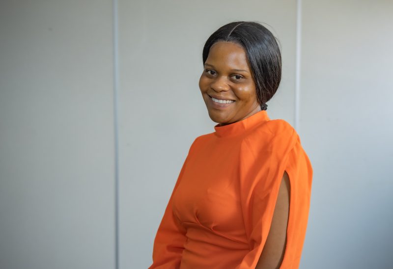 Tapiwa wears an orange dress and is smiling against a white background