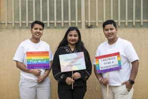 Three members of the Nepal LBTQI+ community stand together holding signs of hope and protest.