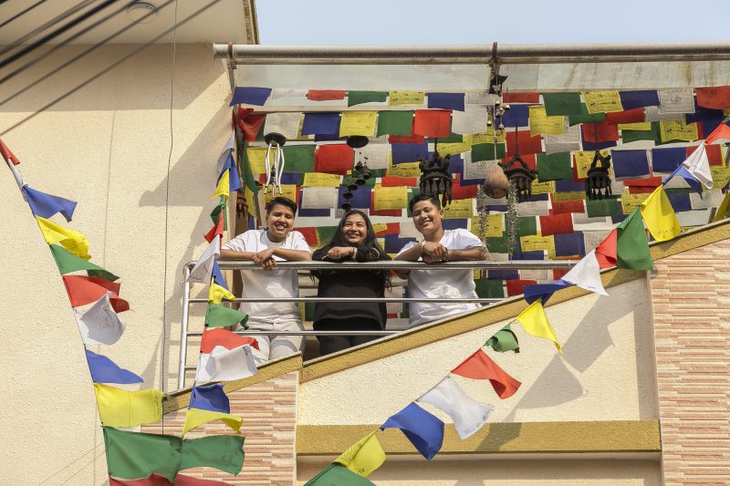 Surrounded by prayer flags, three members of Mitini's community stand smiling at the camera in the sunshine.