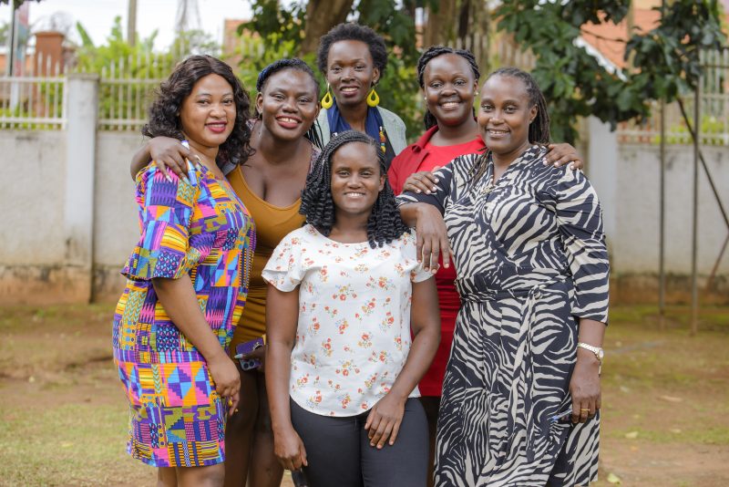 Six women huddle together outside in Uganda. Smiling at the camera.