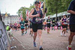 Katie is a white blonde woman, running in black running clothes over the finish line of the London Marathon. Other runners and supporters are in the background.