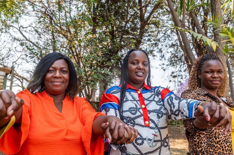 Three women hold hands and look at the camera with trees behind them