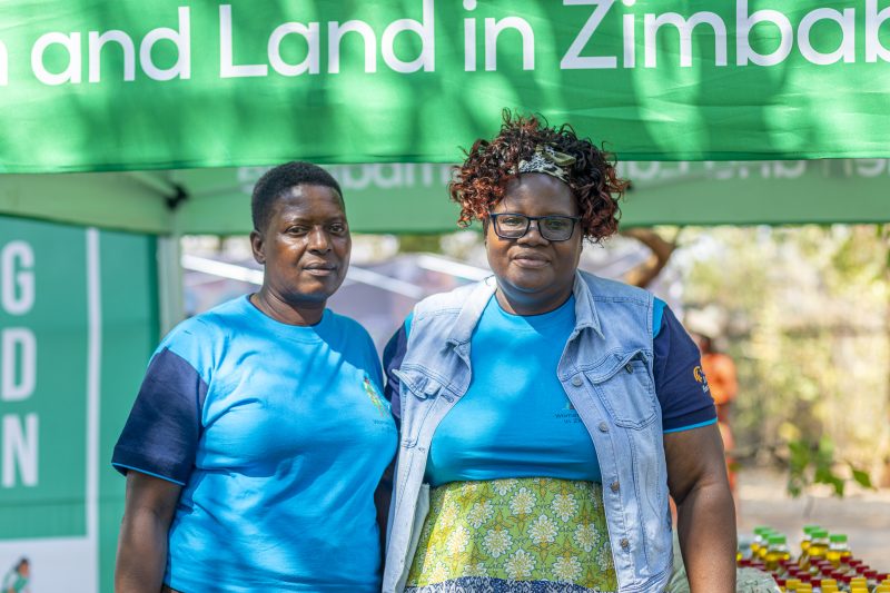 Two women stand in front of the Women and Land in Zimbawbe stall at the Seed Festival both wearing blue Women and Land in Zimbabwe t-shirts