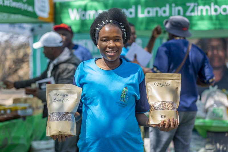 Tinotenda wears a blue Women and Land t-shirt and holds a bag of sugar beans in one hand and a bag of Nyimo in the other.