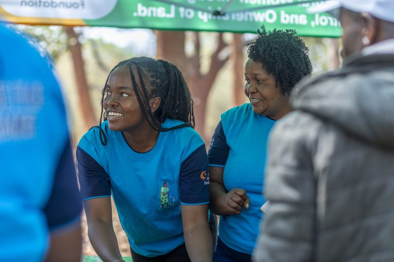Two women wearing blue Women and Land in Zimbabwe t-shirts smile at the stall at the Seed Festival.
