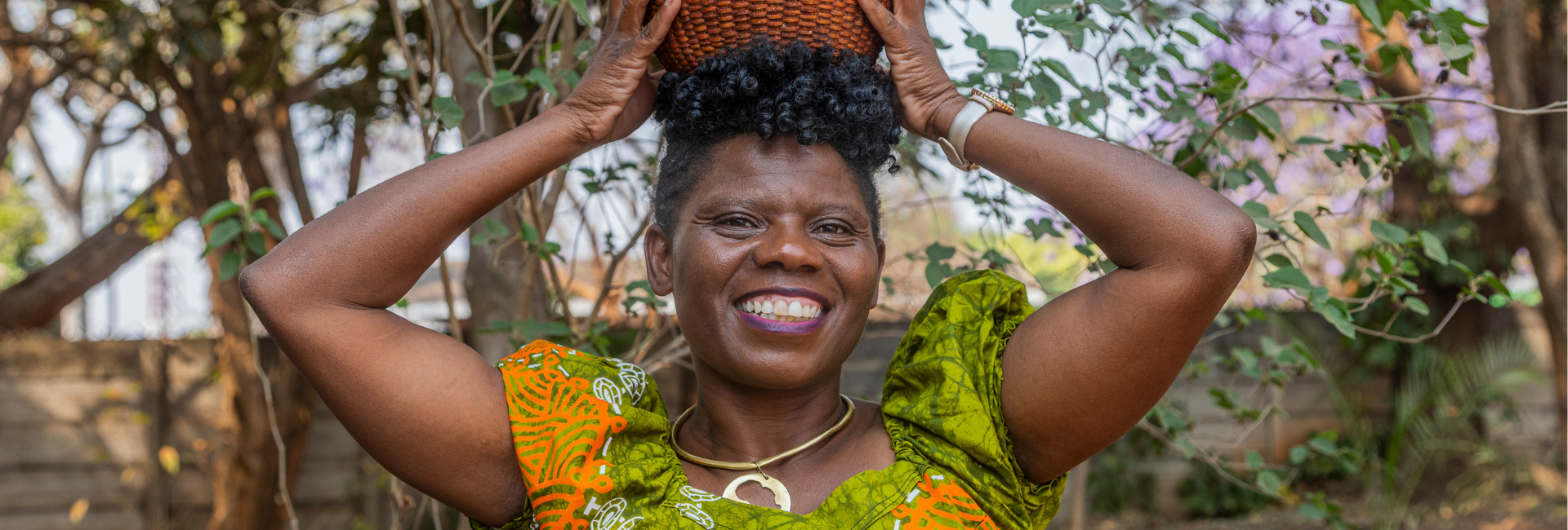 Gloria is part of the Women and Land in Zimbabwe team. She wears a green and orange patterned dress. She is holding a basket on her head and smiling.